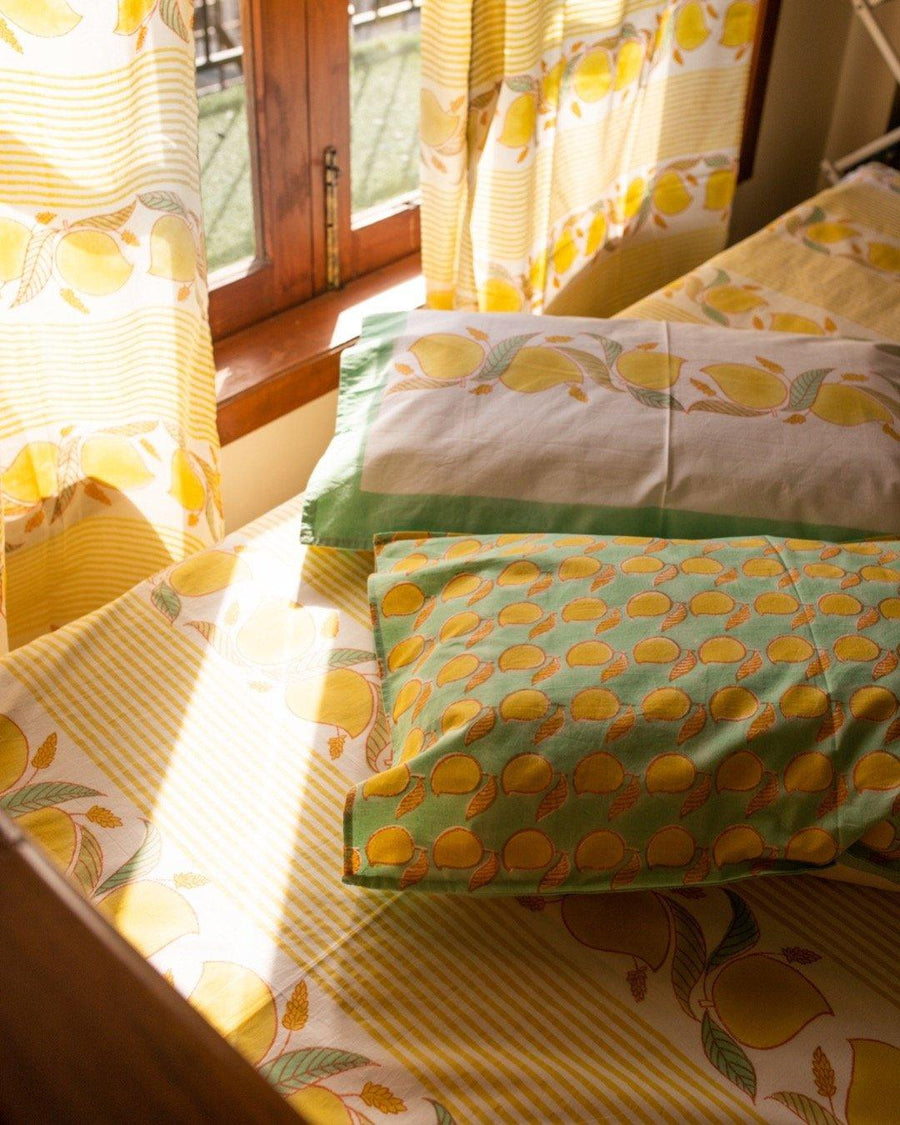 Bedroom set up with yellow block printed Jaipuri bedsheets with mango and vine themed patterns on them, while sunlight streams through the curtains and pillows with similar mango motifs strewn across the bed
